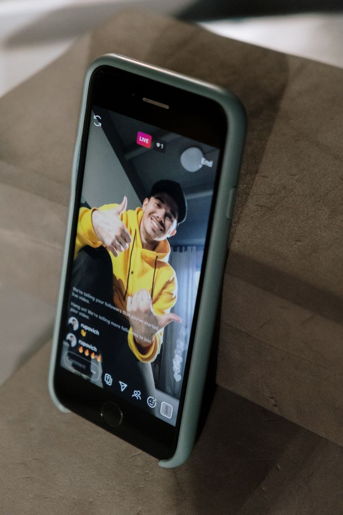 Un homme portant un sweat à capuche jaune anime un direct sur l’écran d’un smartphone, en intérieur.